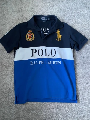 Blå och svart Polo Ralph Lauren Piké - Piké från Polo Ralph Lauren med svart överdel, vit mitt och blå nederdel. Stora broderade loggor och emblem framtill, samt klassisk pikékrage. Tillverkad i bomull och har korta ärmar. Big pony Ralph lauren loga.