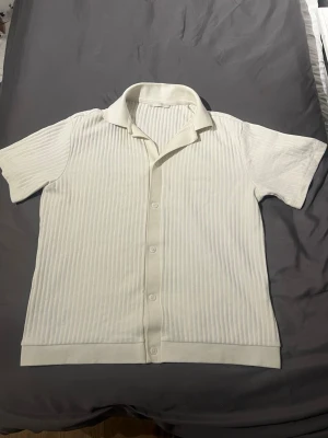 Vit ribbad skjorta från Zara - Kortärmad skjorta från Zara i vit färg med ribbad struktur och klassisk krage. Skjortan har knappar framtill och är tillverkad i ett mjukt material som känns skönt mot huden. Perfekt för en clean och stilren look.