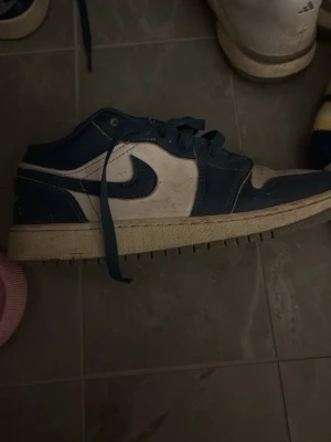 Nike Air Jordan 1 Low blå/vit - Snygga Nike Air Jordan 1 Low sneakers i blått och vitt läder med klassisk swoosh på sidan. Skorna har lågt skaft, blå snörning och en vit mellansula med greppvänlig yttersula. Perfekta för dig som gillar streetwear och sportig stil.