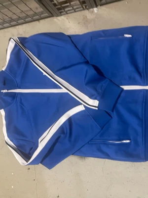 Blå Palm Angels ziptröja med vita ränder - Snygg blå ziptröja från Palm Angels med vita ränder längs ärmarna och vit dragkedja. Tröjan har ståkrage, ribbade muddar och två fickor med dragkedja. Palm Angels-logga tryckt på bröstet. Perfekt för en sportig och trendig look.