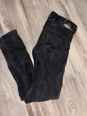 Svarta jeans från Replay - Säljer ett par svarta jeans från Replay med klassisk femficksdesign och diskreta slitningar. Har två hål vid knäna. Jeansen har smal passform och snygga detaljer på bakfickan. Perfekta för dig som gillar en stilren och modern look.
