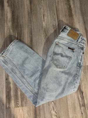Ljusblå raka jeans från Nudie Jeans - Säljer ett par ljusblå jeans från Nudie Jeans med klassisk femficksdesign och raka ben. Jeansen har en snygg tvättad look, läderpatch med logga bak och detaljerade sömmar på bakfickorna. Perfekt för dig som gillar retrostil och avslappnad passform.