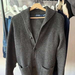 Grå stickad kofta från Polo Ralph Lauren - Mysig grå kofta från Polo Ralph Lauren i grovstickad ull. Koftan har knappar framtill, två fickor och en klassisk krage. Perfekt för lager-på-lager och chill höst- och vinterstil.