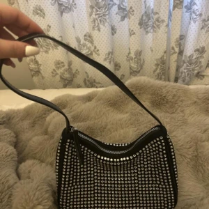 Svart axelväska med glittriga stenar - Snygg svart axelväska täckt med glittrande silverfärgade stenar. Väskan har en enkel axelrem och stängs med dragkedja upptill. Perfekt accessoar för att lyfta vilken outfit som helst med lite extra bling.