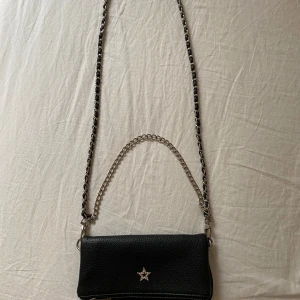 Svart axelväska med kedja och stjärndetalj - Snygg svart axelväska i skinnimitation med silverfärgad kedja och lång axelrem flätad med svart material. Väskan har en glittrig stjärna framtill och dragkedja längs nederkanten. Perfekt storlek för det viktigaste och har ett extra fack på baksidan.