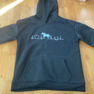 Svart hoodie med hästtryck och fleece - Svart hoodie med vitt tryck av hjärtslag, hjärta, häst och hästsko på bröstet. Mjuk fleece på insidan och stor  framtill. Perfekt för dig som älskar hästar och vill ha något riktigt bekvämt och varmt.  Den är från Temu!!! Står ingen storlek i den men de är storlek S. Väldigt tjock och varm. 