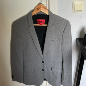 Grå kavaj från Hugo - Stilren grå kavaj från Hugo med klassisk passform. Kavajen har två knappar framtill, fickor med lock och en bröstficka. Insidan är fodrad i svart med snygga röda detaljer. Perfekt för dig som vill ha en clean och modern look.