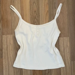 Vit ribbad linnetopp med knappar från brady Melville - Säljer en vit linnetopp med smala axelband och ribbad känsla. Toppen har en enkel design med tre små knappar framtill och är tillverkad i mjuk bomull. Perfekt för varma dagar eller som lager under andra plagg.