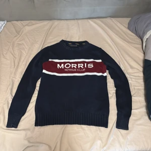 Morris stickad tröja - Snygg stickad tröja från Morris i marinblått med bred vinröd och vit rand över bröstet. Broderad logga framtill, ribbade muddar och klassisk passform. Perfekt för dig som gillar preppy stil och vill sticka ut lite extra.