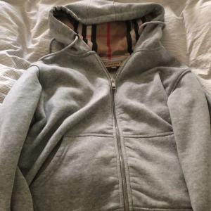 Grå hoodie från Burberry med dragkedja - Säljer en grå hoodie från Burberry med klassiskt rutigt foder i huvan och broderad logga på bröstet. Tröjan har dragkedja framtill, känguruficka och huva med snörning. Perfekt för dig som vill ha en stilren och lyxig vardagslook.