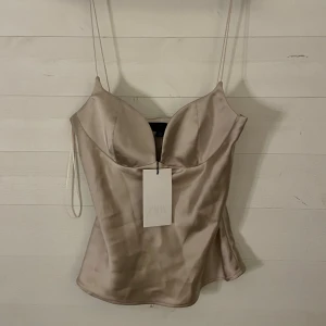 Champagnefärgad satin korsett från Zara - Helt ny med prislappen kvar. Supersnygg champagnefärgad korsettopp från Zara i glansig satin med tunna axelband och markerad byst. Toppen har en figurnära passform och en elegant, lätt skimrande yta som ger en lyxig känsla. Perfekt för att lyfta vilken outfit som helst.