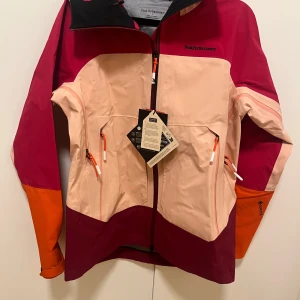 Peak Performance färgstark vindjacka - Snygg vindjacka från Peak Performance i färgblock med ljusrosa, vinröd och orange. Jackan har huva, dragkedja framtill och flera fickor med orange detaljer. Materialet är slitstark polyester och jackan har en sportig vibe med modern passform .Det självklara Gore-Tex C-Knit™-skalet för äventyr året runt ovan trädgränsen. Designat för toppturer med fokus på låg vikt, skydd och ventilation. Utvecklat tillsammans med professionella skidåkare. Ordinariepris är 6500kr.