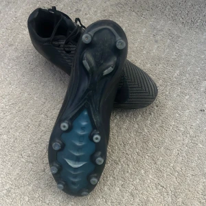 Svarta Nike fotbollsskor med dobbar - Säljer ett par svarta Nike fotbollsskor med snörning och mönstrad ovandel. Skorna har rund tå, platt sula med fasta dobbar och är tillverkade i syntetmaterial. Perfekta för dig som vill ha stilrena och snabba skor på planen.