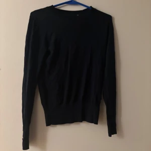 Svart stickad tröja Holly & Whyte - Svart stickad tröja från Holly & Whyte by Lindex. Klassisk modell med rund halsringning och långa ärmar. Snygga guldfärgade knappar vid ärmsluten ger en extra detalj. Perfekt till lager-på-lager under höst och vinter.