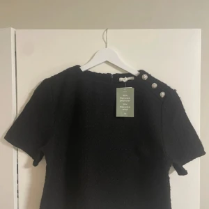 Svart topp med dekorativa knappar H&M - Svart topp från H&M i strukturerat tyg med kort ärm och rund hals. Snygga silvriga knappar med mönster på ena axeln ger en cool detalj. Tillverkad av 90% återvunnen polyester och 10% återvunnen ull. Dragkedja bak för enkel påklädning.