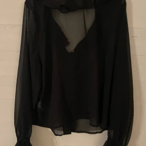 Svart transparent blus med knyt - Svart transparent blus i luftig polyester med v-ringning och volangkant. Långa ärmar med knyt vid handlederna och lös passform. Perfekt för dig som vill ha en stilren men ändå edgy look.