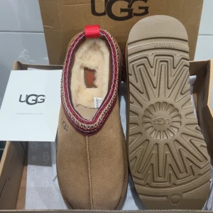 UGG mockatofflor med ullfoder - Bruna UGG tofflor i mocka med mjukt ullfoder och dekorativ kant i rött och beige. Platt sula med UGG-logga på sidan. Perfekta för kalla dagar när du vill ha det extra mysigt. Kan passa i storlek 39/40