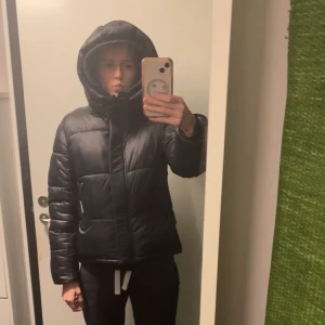 Svart pufferjacka med huva - Intressekoll på min fina vinterjacka från khujo. Köpt för 2 år sedan använd men fortfarande i fint skick. Den är varm och mysig med en väldigt fin luva men funderar på att sälja då jag tycker den är något kort på mig (172cm). Nypris 2049kr kom med bud men inga skambud!