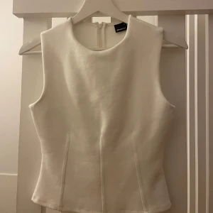 Vit ärmlös topp från Gina Tricot - Säljer en stilren vit topp från Gina Tricot i storlek S. Toppen är ärmlös med rund halsringning och har en figurnära passform. Den är tillverkad i ett mjukt material och har dragkedja bak för enkel påklädning. Perfekt att matcha med jeans eller kjol.