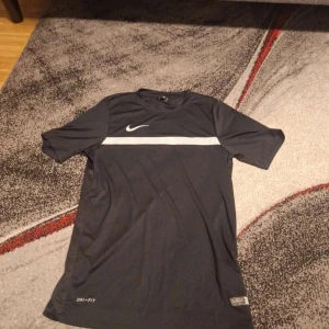 Nike Dri-Fit svart t-shirt - Svart Nike Dri-Fit t-shirt med vit horisontell rand över bröstet och klassisk swoosh-logga. Tillverkad i lätt och ventilerande polyester, perfekt för träning. Kortärmad modell med sportig vibe och normal passform.