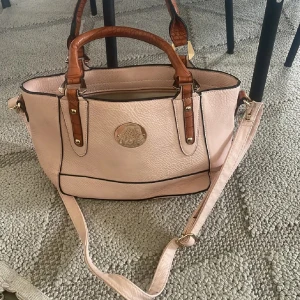 Mulberry ljusrosa handväska med bruna detaljer - Snygg ljusrosa handväska från Mulberry med bruna handtag i croco-imitation och guldfärgade metalldetaljer. Väskan har avtagbar axelrem, svart kantband och ett stort innerfack med dragkedja. Perfekt för dig som vill ha en chic och rymlig väska.