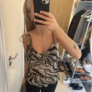 Zebramönstrad linnetopp med knytband - Säljer en snygg zebramönstrad linnetopp med tunna axelband som knyts på axlarna. Toppen har volangdetaljer framtill och en lös passform. Färgerna går i svart, beige och grått. Perfekt för dig som vill sticka ut med ett coolt mönster och öppen rygg.