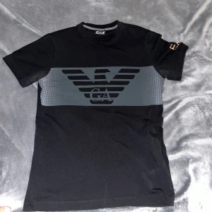 Svart EA7 t-shirt med stor logga - Svart t-shirt från EA7 Emporio Armani med stor grå logga över bröstet och guldfärgad EA7-tryck på ärmen. Klassisk rund hals och korta ärmar. Materialet är mjuk bomull som sitter skönt mot kroppen. Perfekt för dig som gillar stilrena och moderna plagg. Använt fåtal gånger sedan gömd i garderoben. Köpt för 550kr. 