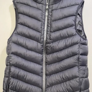 Svart dunväst från Alessandro Zavetti - Snygg svart dunväst från Alessandro Zavetti med quiltad design och dragkedja framtill. Västen har diskret logga på bröstet och bak, samt praktiska fickor med dragkedja. Perfekt för lager-på-lager och stilren look. Storlek S