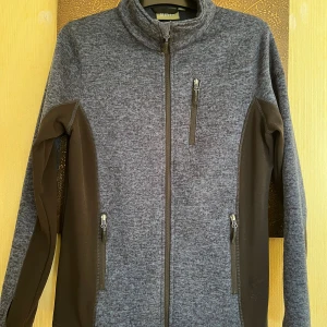 Grå fleecejacka från Strova - Mysig gråmelerad fleecejacka från Strova med svarta paneler på sidor och ärmar. Jackan har hög krage, tre dragkedjeförsedda fickor och hel dragkedja framtill. Perfekt för kyliga dagar och enkel att matcha med det mesta.
