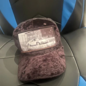 Mörkbrun sammetkeps med biltryck - Säljer en unik mörkbrun keps i sammet med ett coolt tryck av en klassisk bil framtill. Kepsen har en mjuk och lyxig känsla tack vare sammetstyget och passar perfekt för dig som vill sticka ut med din stil.