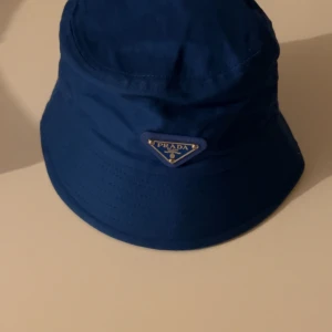 Blå bucket hat  - Snygg blå bucket hat med klassisk trekantig logga framtill i metall. Hatten har en rund form och är tillverkad i ett slätt syntetmaterial som ger en modern vibe. Perfekt för dig som vill sticka ut med en ikonisk accessoar.