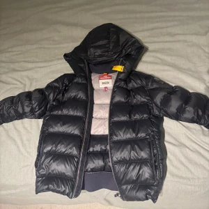 Svart dunjacka Parajumpers Sheen - Svart dunjacka från Parajumpers, modell Sheen, med huva och klassisk gul detalj vid halsen. Jackan har quiltad design, dragkedja framtill och ribbade muddar. Märkespatch på ärmen och två fickor med dragkedja. Perfekt för kalla dagar.