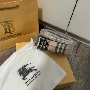 Rutig Burberry bälte med metallspänne - Helt nytt, Snyggt bälte från Burberry med klassiskt rutigt mönster i beige, svart och rött. Bältet har ett stort metallspänne med Burberrys ikoniska monogram. Perfekt accessoar för att lyfta din outfit med en lyxig touch. 