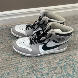Nike Air Jordan 1 High vit/grå 42 - Säljer ett par Nike Air Jordan 1 High i stilren vit och grå färg med svarta detaljer och snörning. Klassisk siluett med svart swoosh och Air Jordan-logga på sidan. Ovandel i skinn och syntet, perforerad tå och högt skaft för ikonisk look.