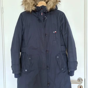 Mörkblå parkas från Tommy Hilfiger - Snygg mörkblå parkas från Tommy Hilfiger med avtagbar huva med fejkpäls, stora fickor och ribbade muddar. Jackan har både dragkedja och knappar framtill samt justerbar midja. Perfekt för kalla vinterdagar. jag köpte i USA, det står storlek XXS men det är mer en XS (jag brukar ha storlek xs/34 från H&M). Den har tecken på slitage men är överlag i fint skick, den är fortfarande lång att bära