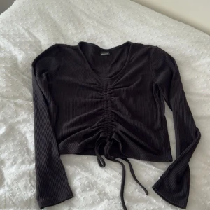 Svart ribbad croppad topp Gina Tricot - Svart långärmad croppad topp från Gina Tricot med ribbad struktur och snygg snörning framtill. Toppen har v-ringning och är tillverkad i mjukt stretchigt material som sitter skönt på kroppen. Perfekt för en trendig och avslappnad look.