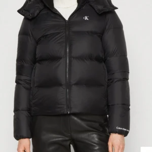Svart pufferjacka Calvin Klein Jeans S - Svart pufferjacka från Calvin Klein Jeans med diskret logga på bröstet och ärmen. Jackan har dragkedja framtill, hög krage och avtagbar huva. Tillverkad i glansig polyester och har en klassisk, boxig passform som är både varm och snygg till vintern. Använd få gånger. Köptes för 2000kr men säljer för 600kr