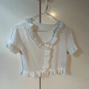 Vit volangblus från SHEIN - Supersöt vit blus från SHEIN i storlek S. Toppen är croppad med korta ärmar, volangdetaljer längs krage, ärmslut och nederkant samt knappar framtill. Perfekt för en trendig och feminin look.