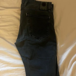 Replay Anbass jeans i mörkgrå denim - Säljer ett par snygga Replay Anbass jeans i mörkgrå tvätt. Klassisk femficksmodell med Replay-logga vid fickan och broderad detalj på insidan av linningen. Jeansen har en modern passform och är tillverkade i mjuk denim med lite stretch.