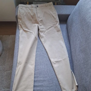 Beiga chinos från H&M, stl 164 - Snygga beiga chinos från H&M i storlek 164. Byxorna har klassisk rak passform, sidfickor och bälteshällor. Tillverkade i bomullsmix med 20% återvunnet material. Perfekta för en clean och stilren look. Helt oanvända, lappen sitter kvar till och med. Nypris 200 kr