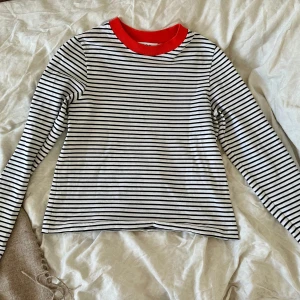 Randig långärmad tröja från COS nyskick - Säljer en snygg långärmad tröja från COS i vitt med svarta tunna ränder och en cool röd krage. Tröjan är i mjuk bomull och har en klassisk, rak passform. Perfekt för dig som gillar stilrena och trendiga basplagg.