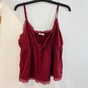 Vinröd linnetopp med spets från Nelly - Säljer en vinröd linnetopp från Nelly med tunna axelband och v-ringning. Toppen har fina spetsdetaljer längs kanterna och en liten rosett framtill. 