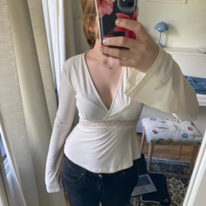 Vit festtopp med spetsdetalj - Supersnygg vit omlottopp med djup v-ringning och långa ärmar. Toppen har en bred spetskant i midjan som ger en romantisk vibe och markerar midjan snyggt. Materialet är stretchigt och lätt, perfekt för att skapa en feminin siluett.