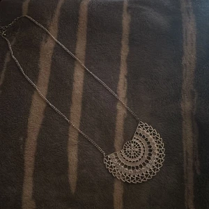 Statement halsband i silver med stenar - Unikt statement halsband i silverfärg med halvcirkelformad, geometrisk hängare. Hängaren har ett detaljerat mönster av cirklar och trianglar, dekorerat med gnistrande stenar som ger en glittrig effekt. Perfekt för att lyfta vilken outfit som helst.