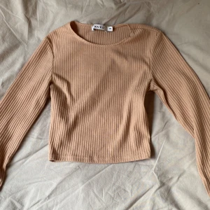 Beige ribbad långärmad topp NA-KD - Säljer en beige ribbad långärmad topp från NA-KD i storlek XXS. Toppen har en rund halsringning och är croppad i modellen. Perfekt att styla med jeans eller kjol för en enkel och snygg look.