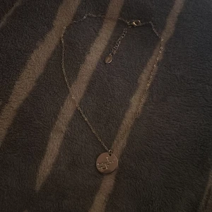 Guldigt halsband med stjärntecken - Snyggt halsband i guldton med tunn kedja och rund berlock. Berlocken har ett stjärnteckenmönster dekorerat med små glittrande stenar. Perfekt accessoar för dig som gillar personliga smycken och astrologi.