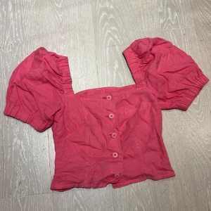 Rosa blus med puffärmar och knappar - Supersöt rosa blus med puffiga korta ärmar och knäppning framtill. Toppen har fyrkantig urringning och är croppad i modellen. Det står tyvärr inte vilken storlek det är eller var den är ifrån, men skulle gissa på en lite större M🤗