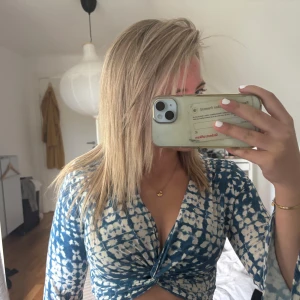 Blåvit croppad omlottopp med mönster - Säljer en croppad omlottopp i blått och vitt med unikt mönster. Toppen har långa ärmar, v-ringning och twistad detalj framtill. Resår i midjan baktill för skön passform. Perfekt för dig som gillar trendiga och coola plagg med lite edge.