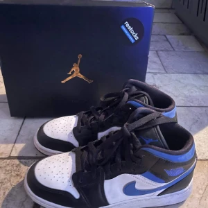 Nike Air Jordan 1 Mid svart/blå/vit - Säljer ett par Nike Air Jordan 1 Mid med svart, blå och vit färgkombination. Klassisk siluett med svart snörning, blå swoosh och detaljer samt perforerad tå. Skorna har en hög modell och är tillverkade i skinn och syntet. Perfekta för dig som gillar streetwear och sneakers.