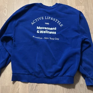 Blå sweatshirt Movement & Wellness - Blå sweatshirt med vit text på bröstet och ryggen där det står 'Movement & Wellness' och 'Active Lifestyle'. Tröjan har rund halsringning, ribbade muddar och är tillverkad i mjuk bomull. Perfekt för en sportig och avslappnad look.